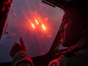 Ultra-realistic editorial photo capturing a mid-shot from inside a Philippine patrol plane as three bright red signal flares streak upward outside the window near Subi Reef. The pilot’s tense silhouette is visible in the foreground, hand on controls, illuminated by the orange flare glow. Outside, blurred shapes of distant Chinese vessels and haze over the disputed waters. Candid, high-emotion moment frozen mid-action, slight motion blur on flares, subtle lens flare and grainy ISO 1600 texture. Background slightly out of focus to emphasize the flare trajectory and rising tension in the South China Sea.